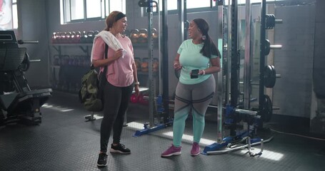 Diverse females talking at gym rack, teal showing phone, pink fixing duffel,towel prepping workout