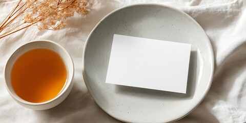 Overhead shot of a blank postcard mockup placed on a simple, white plate. A single, beautifully crafted cup of clear tea is nearby