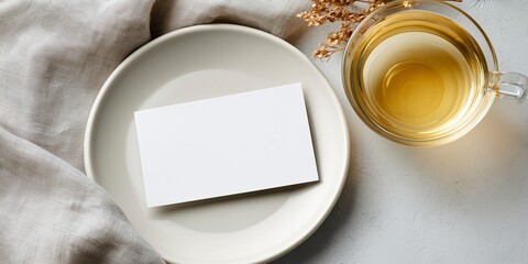 Overhead shot of a blank postcard mockup placed on a simple, white plate. A single, beautifully crafted cup of clear tea is nearby