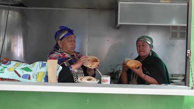 Two mapuche women in traditional clothing enjoying a meal of sopaipillas and mate in a food stall