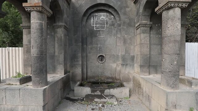 Zoom out revealing a traditional Armenian stone drinking fountain known as Pulpulak. The grey volcanic tuff structure features arches and columns situated outdoors on a sunny day.

