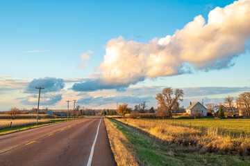 A paved rural road runs through open farmland with distant farm buildings and utility poles visible, surrounded by fields during autumn in Ontario.