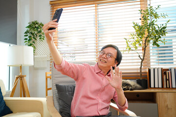 Elderly man smiling while taking selfie with smartphone. Cozy living room setting with plants and soft furnishings. Concept of social connection, technology