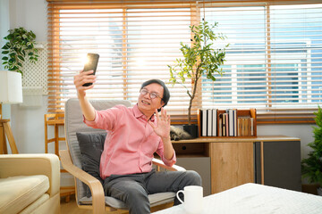 Elderly man smiling while taking selfie with smartphone. Cozy living room setting with plants and soft furnishings. Concept of social connection, technology
