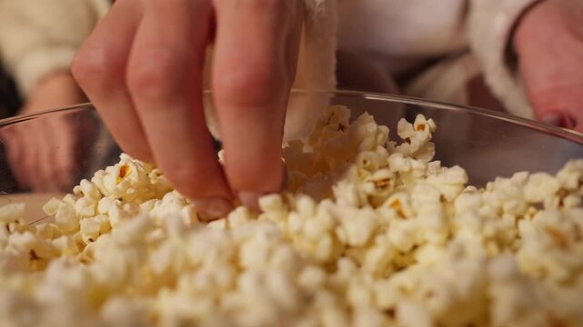 Closeup of mother and daughter hands taking popcorn from large glass bowl while enjoy movie night together, representing moment of family bonding and cozy quality time at home