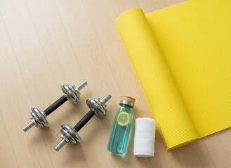 Fitness equipment including dumbbells, a water bottle with lemon, and a towel placed beside a yellow yoga mat, perfect for workouts, exercise routines, and healthy living.
