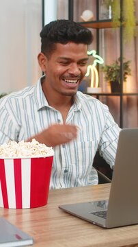Indian man at home watches comedy on laptop eats popcorn and laughs enjoys movie time and relax. Arabian guy at desk holds cup of popcorn smiles looks at screen leans back and feels joy fun nice mood.