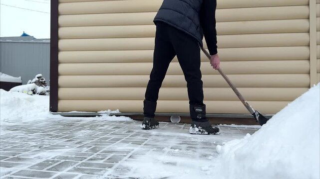 Individual in black winter attire shovels snow from a paved surface beside a building, demonstrating snow removal techniques and progress in clearing the area