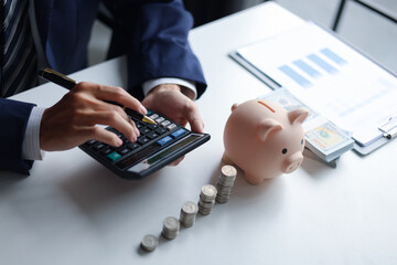 A person using a calculator with a piggy bank, stacks of coins, and US dollar bills on a table. Concept of saving money and financial planning.