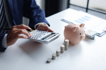 A person using a calculator with a piggy bank, stacks of coins, and US dollar bills on a table. Concept of saving money and financial planning.