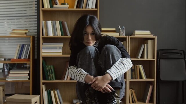 Portrait of thoughtful teenage girl with long black hair wearing emo outfit sitting on chair in college psychotherapists office facing camera with frustrated expression