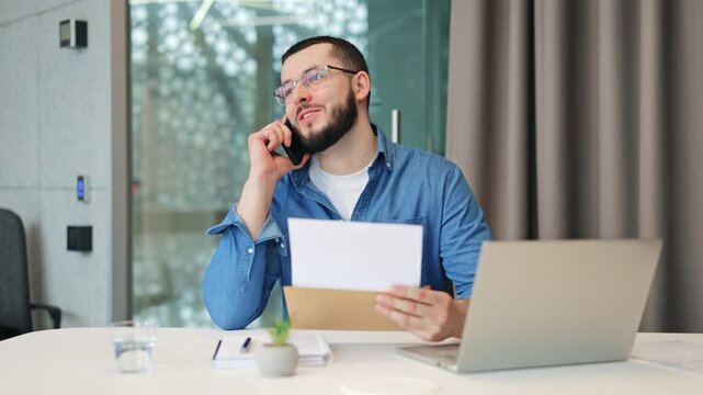 Professional smiles, speaks on mobile, reviewing important documents. Joy and satisfaction are evident from receiving positive news while working in modern office environment, successful communication