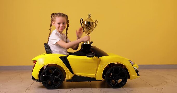 A little girl drives a yellow car, proudly shows a winner&rsquo;s gold trophy to the camera, and waves her hand against a bright orange background. This vibrant 4K video symbolizes success, achievement, vic