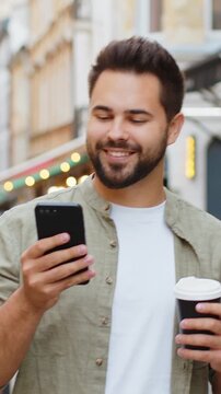 Happy smiling man use smartphone typing text messages browsing internet social media web app working chatting online enjoying morning to-go coffee hot drink outdoor. Boy tourist walking in city street