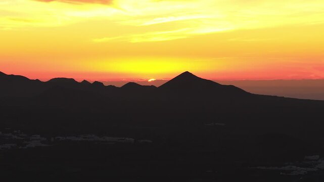 Elevated view shows the sun sinking behind a triangular cone and cratered peaks in Lanzarote, Canary Islands. Warm gradient backlights ridgelines and faint village buildings.