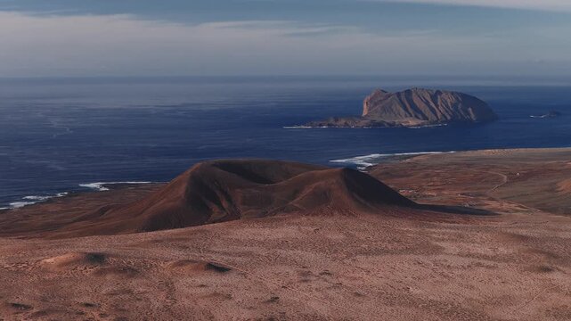 Soft daylight aerial pan and flyover show volcanic plains, twin cinder cones, ridgelines, crater rims, and dirt tracks with La Graciosa and islets off Lanzarote.