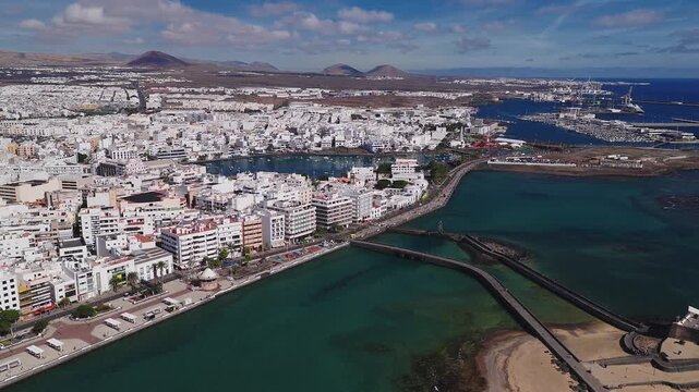 Aerial view of Arrecife, Lanzarote shows waterfront, promenade, Castillo de San Gabriel with twin bridges, Charco de San Gines, marina, port cranes, and volcanic cones.