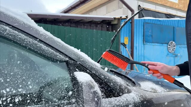 Individual using a snow brush to clear snow from the windshield and roof of a car, showcasing the action of snow removal in a winter setting with visible snow accumulation