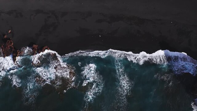 Aerial top down view shows deep teal swells and white foam on black volcanic sand in Lanzarote, Canary Islands, with rust colored lava rock outcrops along the shore.