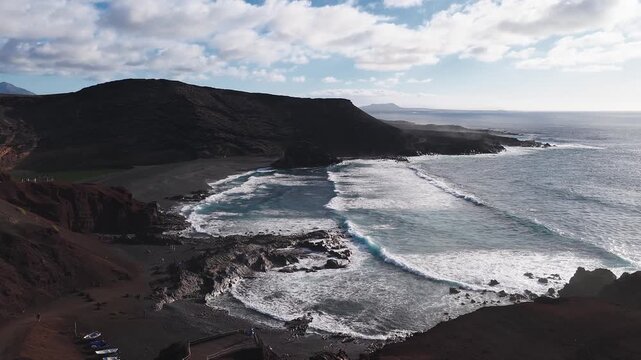 Aerial scene of El Golfo in Lanzarote shows Charco Verde lagoon, black sand, red cliffs, turquoise surf, rolling swells, white lined waves, boardwalk figures, and boats.
