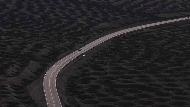 Aerial view of La Geria vineyards in Lanzarote, Canary Islands. A car, another vehicle, and walkers move along a sinuous two lane road across black volcanic soil.