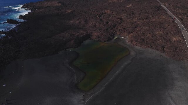 Aerial view of El Charco de los Clicos on Lanzarote, a green lagoon by black volcanic sand and lava fields. Waves break, cars move on a coastal road, tiny figures walk.