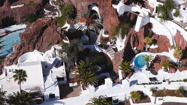 Aerial view of LagOmar in Lanzarote shows white curved buildings, caves, staircases, terraces, palm trees, black gardens, turquoise pools, and visitors in midday sun.