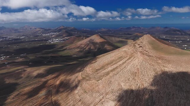 Aerial view of Lanzarote ridgeline, conical craters, Atlantic horizon, white houses, fields, and moving clouds over ochre slopes under high contrast light.