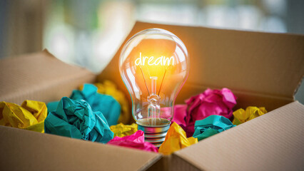Glowing light bulb with word dream inside placed in cardboard box full of colorful crumpled paper creative concept symbolizing ideas innovation imagination and thinking outside the box
