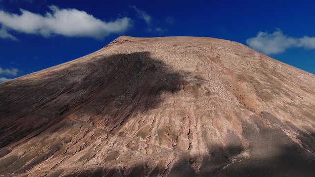 Aerial view of a barren volcanic cone in Lanzarote, Canary Islands. Steep rippled slopes, lava gullies, switchback trail, and moving cloud shadows in bright daylight.