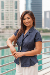 Asian businesswoman stands confidently on a railing in a smart city, showcasing her professional style while working outdoors in a busy setting.