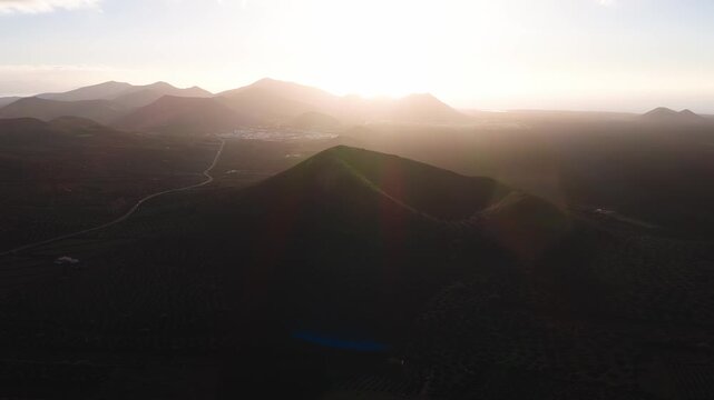 Aerial view shows a cinder cone, layered volcanic peaks, and coastline near a small white settlement in Lanzarote. A road winds through dark lava and farm plots.