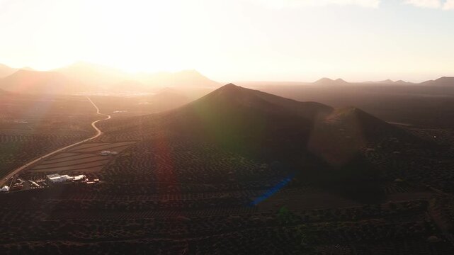 Aerial camera glides and pans over Lanzarote, Canary Islands, La Geria pits, cinder cone, road, white farmhouses, distant mountains, sea, golden hour backlight.