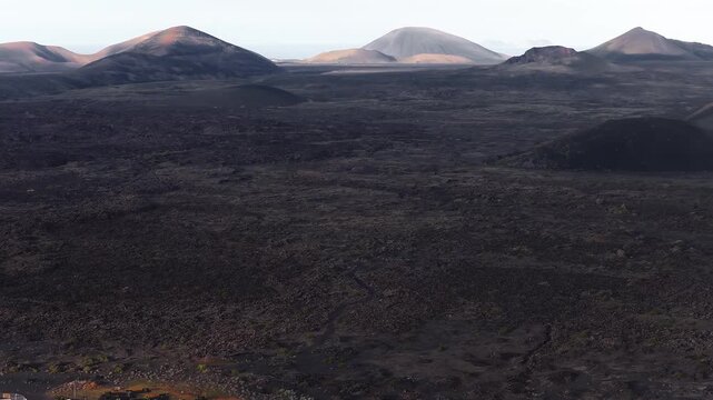 High, slow aerial movement reveals dark lava, cinder cones, shallow craters in Lanzarote. Golden hour side light casts long shadows, Atlantic on the horizon.