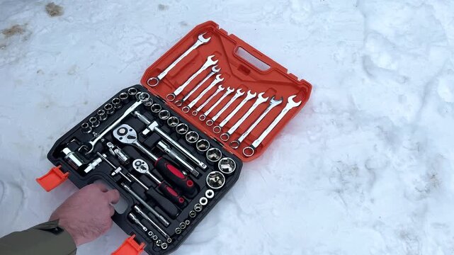 Hands open an orange tool case on snow, revealing a neatly organized set of tools including wrenches and sockets, showcasing preparation for mechanical work outdoors