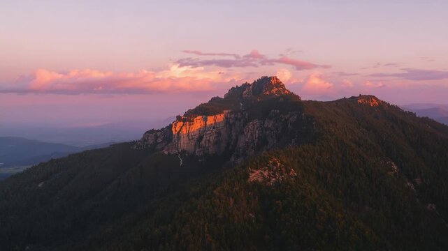 Pulling camera closing to rocky mountain summit, sunlight brightening cliff face and clouds at dawn