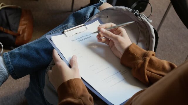 Over shoulder shot of teenage college student rolling pen between fingers while filling in registration form for mental health first aid course in counseling room