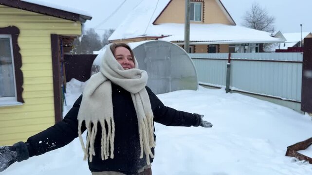 Young woman in winter attire joyfully plays in the snow, preparing to throw a snowball while surrounded by a snowy landscape and a cozy house in the background