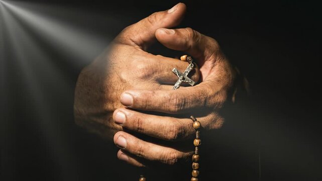 Mans hands clasping a rosary with cross, bathed in divine light. Religious prayer and catholic devotion footage.