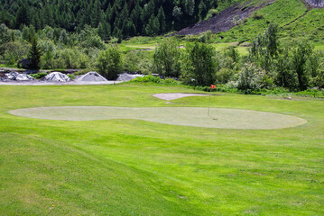 Golf green with flag on a mountain course surrounded by lush forest and alpine landscape in Courmayeur on a sunny summer day.