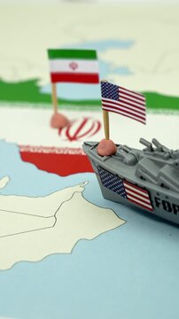 Conceptual representation of the geopolitical conflict between the United States and Iran, showing a toy warship with an American flag sailing towards a map of the Iranian coast