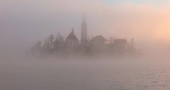 Bled am Morgen zur goldenen Stunde, Nebelschwaden ziehen &uuml;ber die Landschaft und den See, ruhige Morgenstimmung mit warmem Licht und klarer Staffelung von Wasser, Ufer und Hintergrund