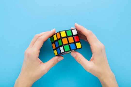 Young adult woman hand fingers holding and playing with colorful plastic rubik cube on light blue table background. Pastel color. Closeup. Point of view shot. Top down view.