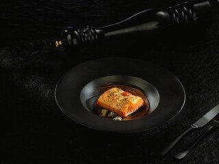 A beautifully presented salmon fillet sits on a sleek black plate, topped with fragrant herbs and...