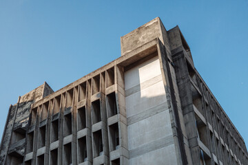A low-angle view of a massive, weathered Brutalist concrete building facade featuring a repetitive geometric pattern of vertical fins and deep shadows set against a clear blue sky.