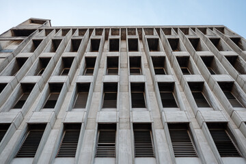 A low angle architectural view of a weathered grey concrete building facade featuring a repetitive geometric grid of rectangular windows with metal louvers.