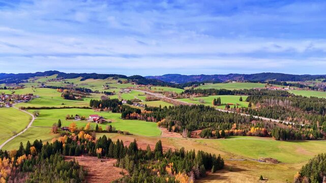 Rolling hills and patchy forests stretch across the landscape. Fields of green grass are interspersed with trees displaying autumn colors.