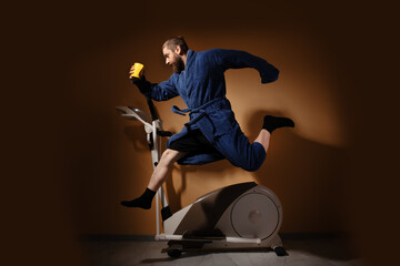 Man jumps on stationary bike in bathrobe, holding coffee cup, against brown background. Concept of multitasking morning routine.