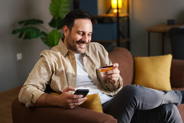 Smiling man making online payment with smartphone and credit card while relaxing on sofa
