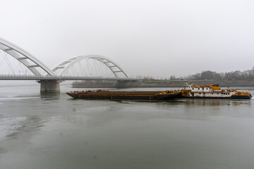Obraz premium Cargo barge sailing on a wide river beneath a modern arched bridge on a foggy day. Industrial transport, calm water, muted tones, overcast weather, quiet atmosphere